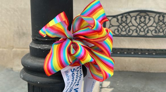 Pride ribbon tied to a street light outside near a bench.