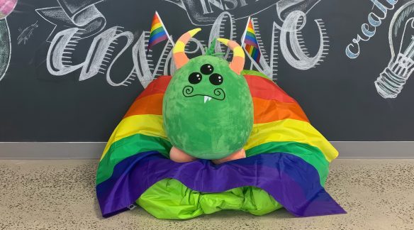The 3i monster holding Pride flags on a bean bag chair.